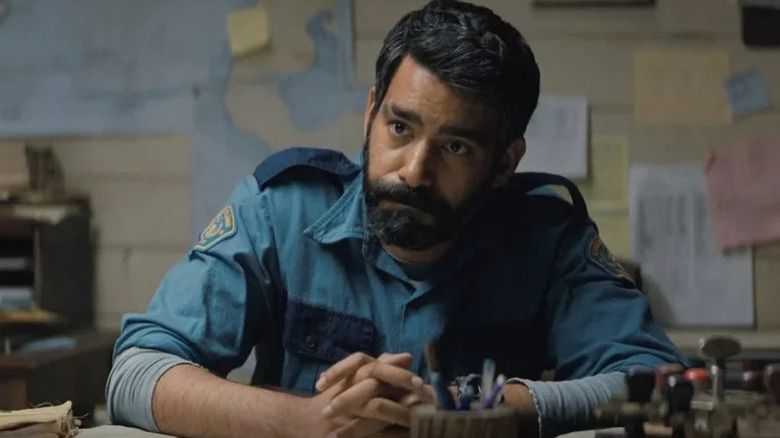 Rahul Kohli sitting at desk in blue police uniform in Midnight Mass