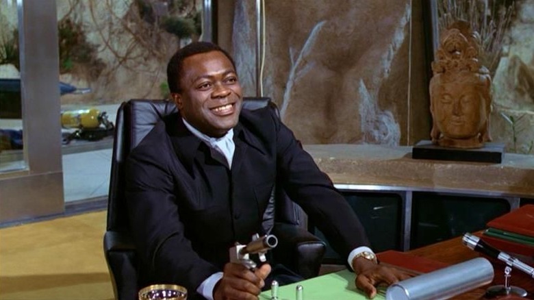 Yaphet Kotto as Mr. Kananga sitting behind a desk, smiling, holding a gun in Live and Let Die.