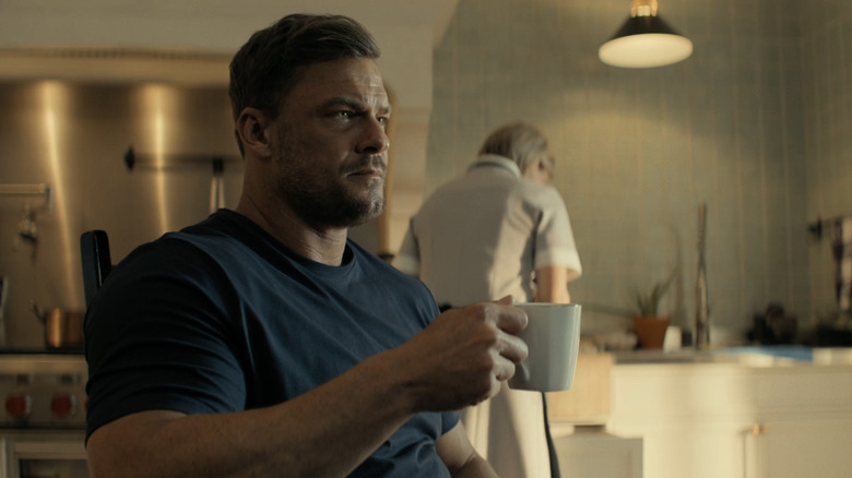 Alan Ritchson's Jack Reacher holds a coffee mug as he sits at a kitchen table in Reacher