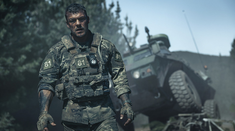 Soldier 81 standing in fatigues with a military vehicle behind him in War Machine