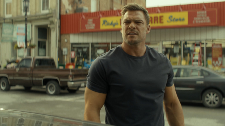 Alan Ritchson as Jack Reacher stands beside a car in a street in Reacher