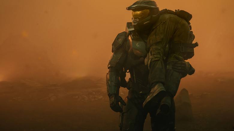 Pablo Schreiber as Master Chief carrying a wounded soldier over his shoulder in a landscape filled with orange haze in Halo