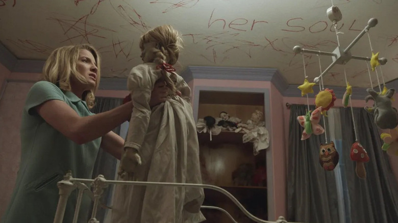 Mia holds up Annabelle the doll in a bedroom with creepy words scrawled on the ceiling in Annabelle