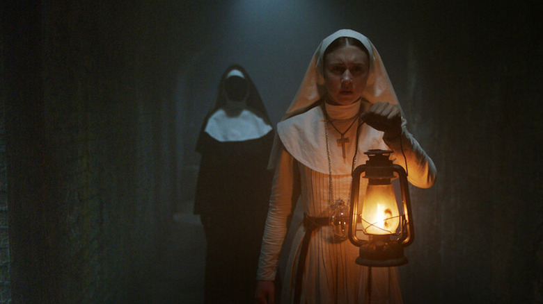Sister Irene holds up a lantern in a dark hallway while the Nun creeps up behind her in The Nun