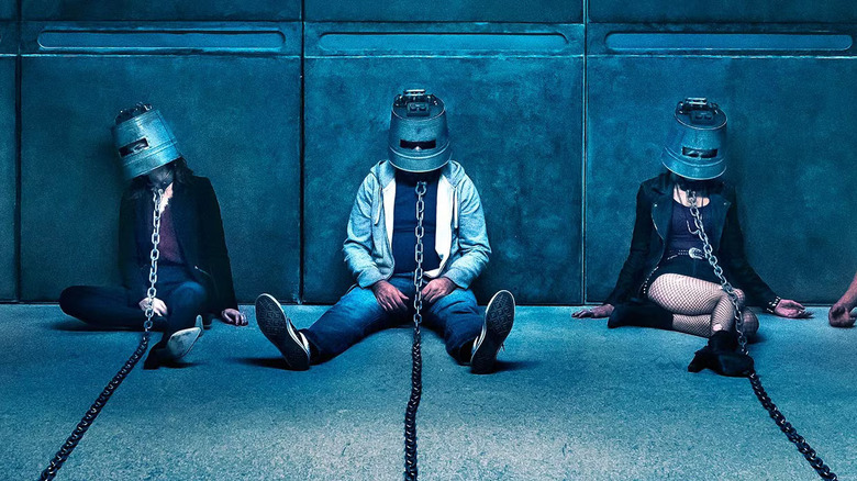Three people laying on the floor against a wall with buckets over their heads and chains around their necks leading offscreen in Jigsaw
