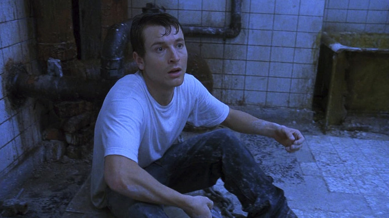 Leigh Whannell as Adam Stanheight sitting on the bathroom floor next to some pipes in Saw