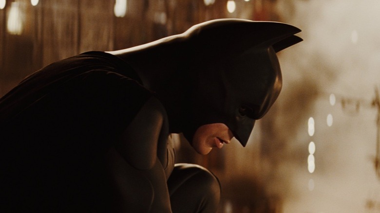 A side-profile of Christian Bale as Bruce Wayne/Batman, staring down out of frame with a smoky city populating the background.