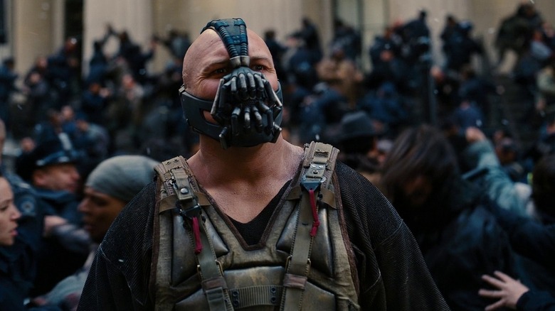 Tom Hardy as Bane in The Dark Knight Rises, looking out of frame toward the middle distance with a chaotic mob behind him.
