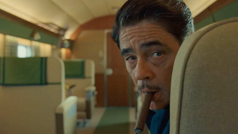 Benicio del Toro as Anatole "Zsa-Zsa" Korda, smoking a cigar and looking over a seatback in The Phonecian Scheme