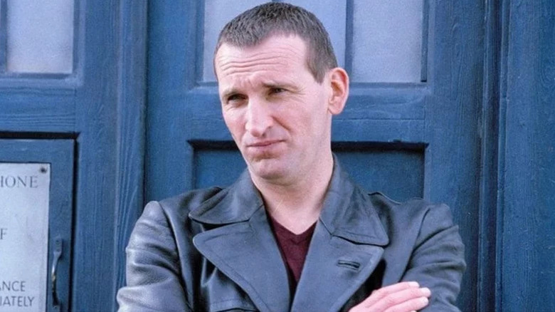 The Ninth Doctor (Christopher Eccleston) stands nonplussed with his arms crossed by the TARDIS in Doctor Who