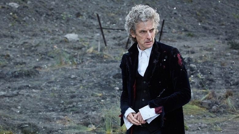 The Twelfth Doctor (Peter Capaldi) stands outside wearing a red velvet jacket as it snows in Doctor Who
