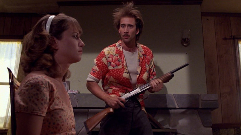 Nicolas Cage as Herbert holding a gun and and Holly Hunter as Edwina McDunnoughin sitting in a chair in front of him in Raising Arizona