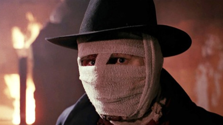 Liam Neeson as Peyton Westlake AKA Darkman wearing a hat with his face wraped in bandages in front of a fire in Darkman