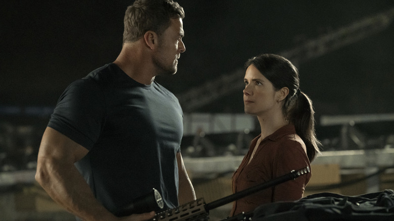 Alan Ritchson as Jack Reacher standing next to Sonya Cassidy as Susan Duffy in front of a table full of guns in Reacher