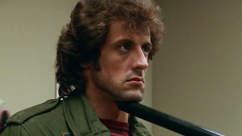 Sylvester Stallone's John Rambo looks across frame as someone off screen places a truncheon under his chin in First Blood