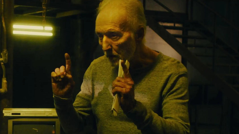 A focused-looking John Kramer (Tobin Bell) points both of his index fingers while holding a rolled-up paper, explaining something in a dimly lit, industrial-looking room in Saw X