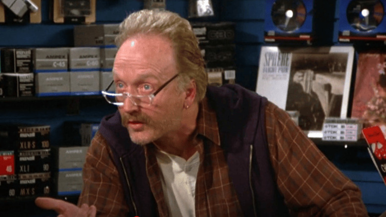 Ron (Tobin Bell), wearing glasses and a plaid shirt, leans forward on the counter with video tape stock and records behind him in a scene from Seinfeld