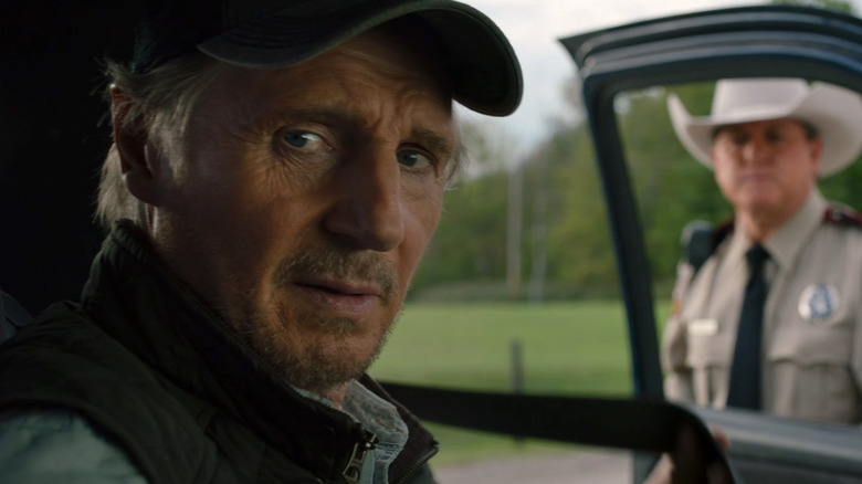 Liam Neeson's Jim Hanson sits in the driver's seat of his car while a police officer stands at his window in The Marksman