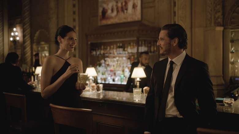 Eva Victor as Nadia Hynes holding a drink by a bar and standing next to Pedro Pascal as Alex Serian in Behemoth!