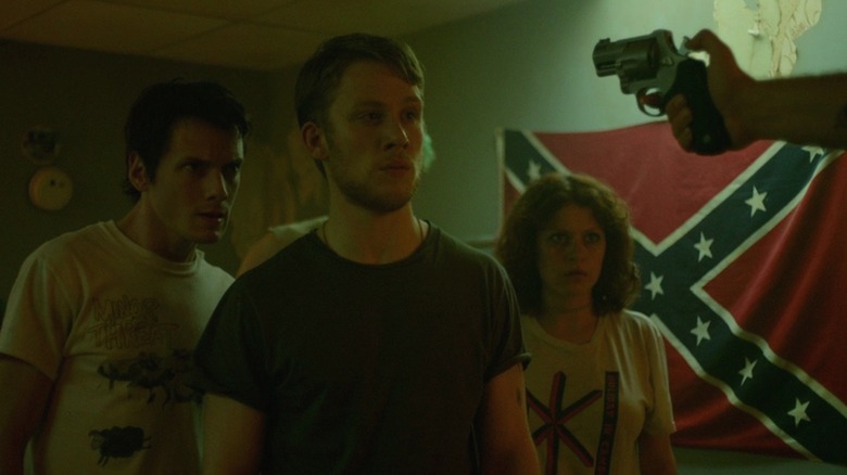 A gun pointed at Anton Yelchin as Pat, Joe Cole as Reece, and Alia Shawkat as Sam in Green Room, in front of a confederate flag