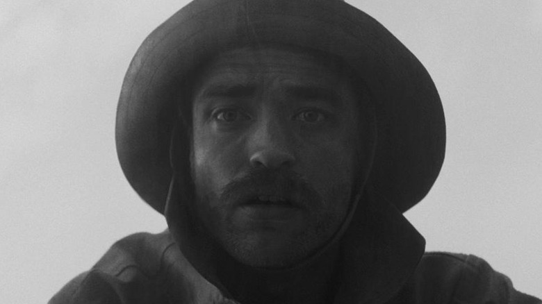 a black and white photo of Robert Pattinson as Thomas in The Lighthouse, wearing a hat, staring down at the camera in awe