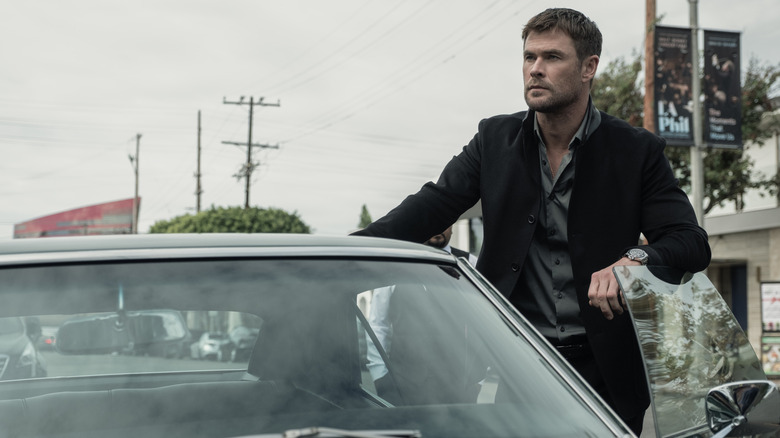 Chris Hemsworth as Mike Davis wearing a suit jacket and leaning on the door to his car in Crime 101