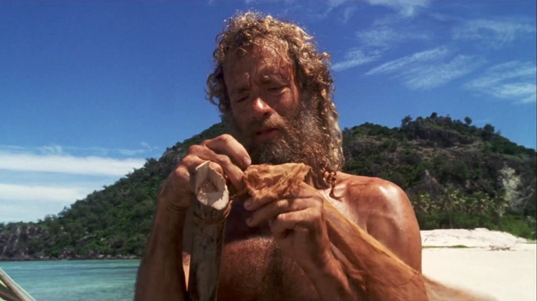 Tom Hanks as Chuck Noland in Cast Away, backdropped by the island, dirty and bearded as he works to maintain his shelter.