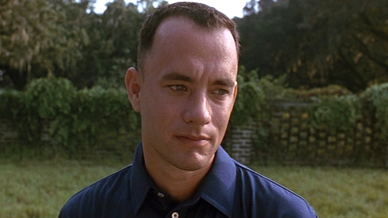 Tom Hanks as Forrest Gump in Forrest Gump, standing in front of a grassy wall and looking off toward the side of the frame.