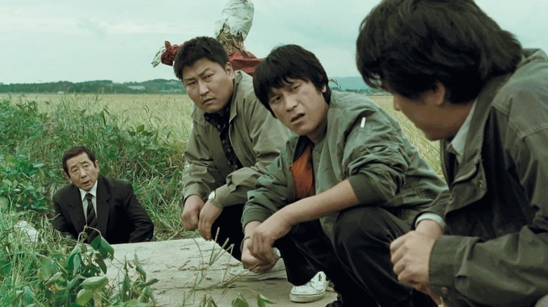 Park Doo-man (Song Kang-ho) and Seo Tae-yoon (Kim Sang-kyung) crouch while talking to police detectives at a crime scene in Memories of Murder