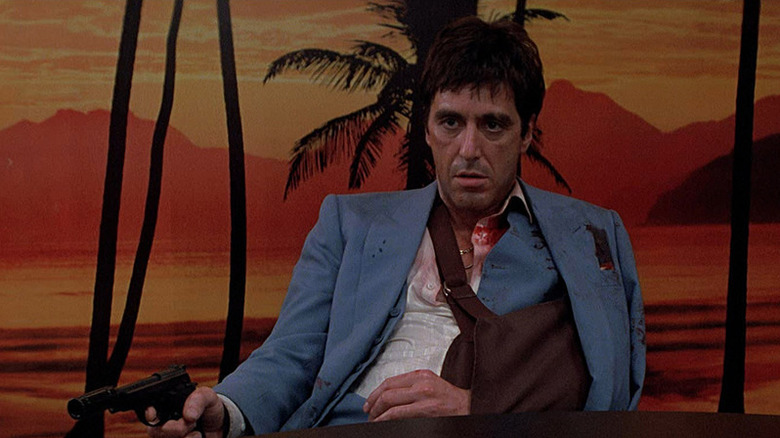 A wounded Tony Montana (Al Pacino) sits at a table holding a pistol with his arm in a sling in Scarface (1983)
