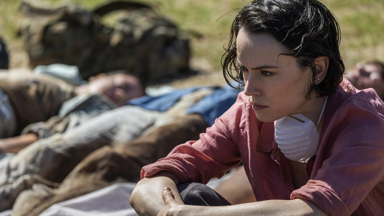 Ava sits in front of a line of dead bodies with a mask dangling around her neck in We Bury the Dead