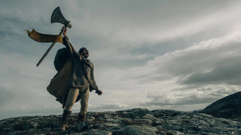 Sir Gawain (Dev Patel) holding axe raised to the sky in mountainous landscape