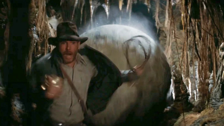 Harrison Ford as Indiana Jones holding gold object and running from massive boulder behind him in Indiana Jones and the Temple of Doom