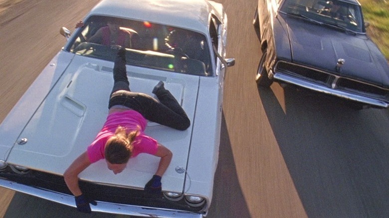 Zoe Bell in pink shirt and gloves laying on hood of white car with dark car behind in Death Proof