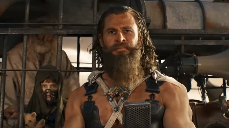 Chris Hemsworth as Dementus in Furiosa: A Mad Max Saga, shirtless but wearing a vest to hold his weaponry, looking forward with Alyla Browne as young Furiosa caged behind him.