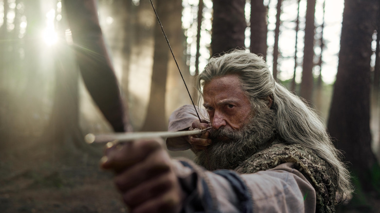Hugh Jackman as Robin Hood holding a bow and arrow in a sunlit forest in The Death of Robin Hood