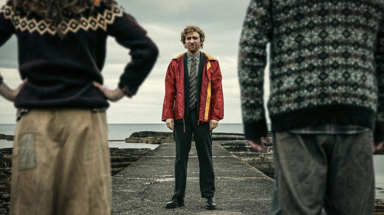 Daniel standing on a dock facing the camera, looking at Isla and Sandy in The Incomer