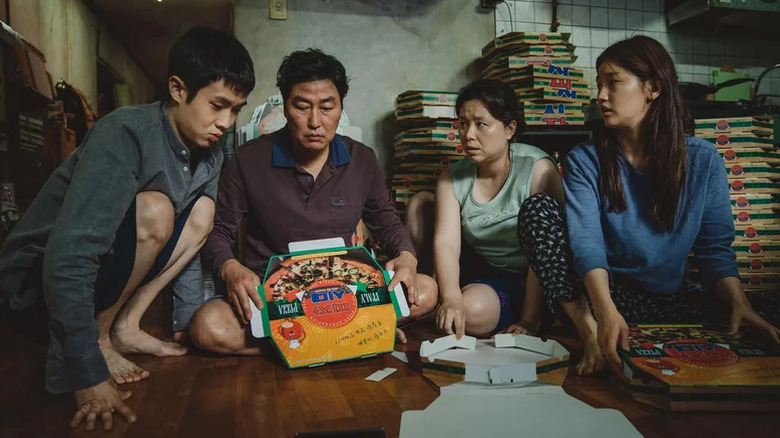 Choi Woo-shik as Kim Ki-woo, Song Kang-ho as Kim Ki-taek, Jang Hye-jin as Chung-sook, and Park So-dam as Kim Ki-jung sitting on the floor surrounded by pizza boxes in Parasite