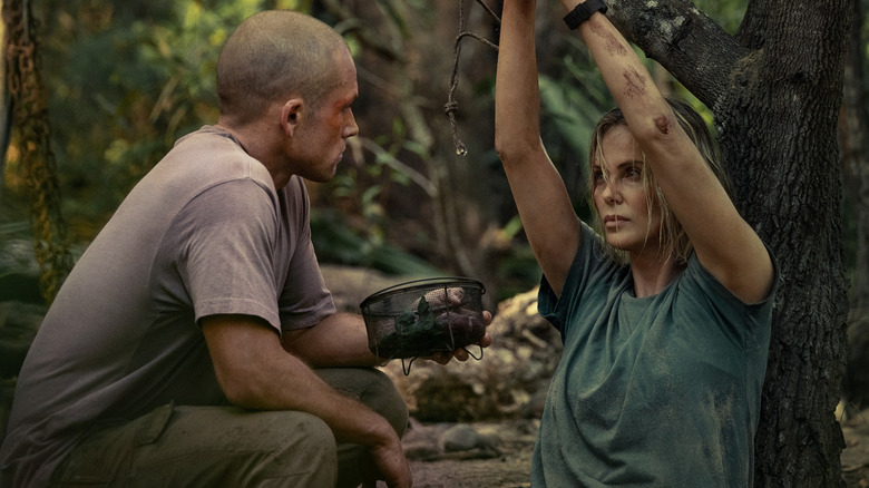Taron Egerton as Ben crouching in front of Charlize Theron as Sasha with her hands tied to a tree branch in Apex