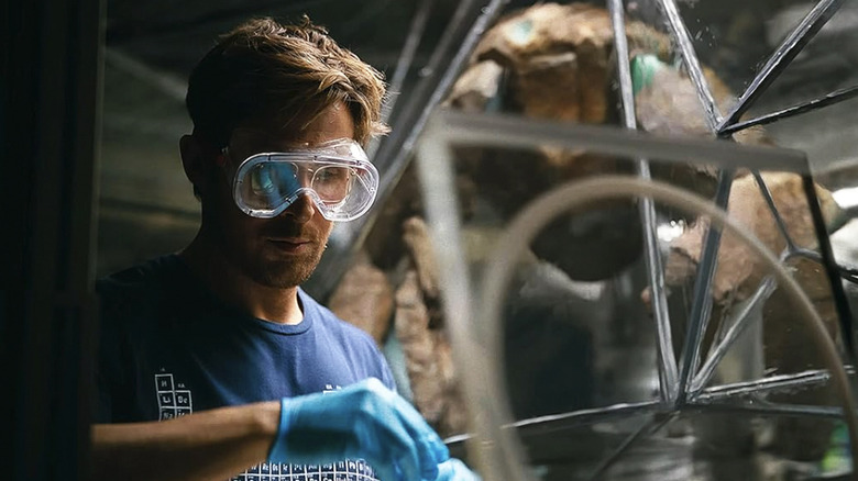 A goggles-clad Ryland Grace does a science experiment while Rocky watches inside his containment material in Project Hail Mary