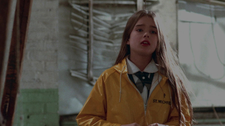 Paula E. Sheppard as Alice Spages in Alice, Sweet Alice, standing among a decrepit building in a yellow coat as she looks on past the frame.