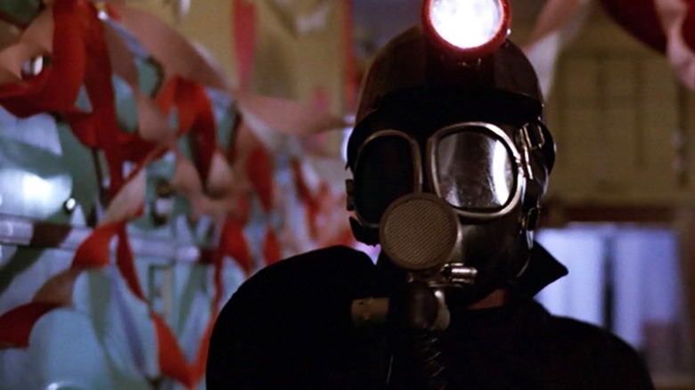 Peter Cowper as The Miner in My Bloody Valentine, dressed in his signature suit and gas mask, standing in close focus against a background of a building strewn with banners on the walls.
