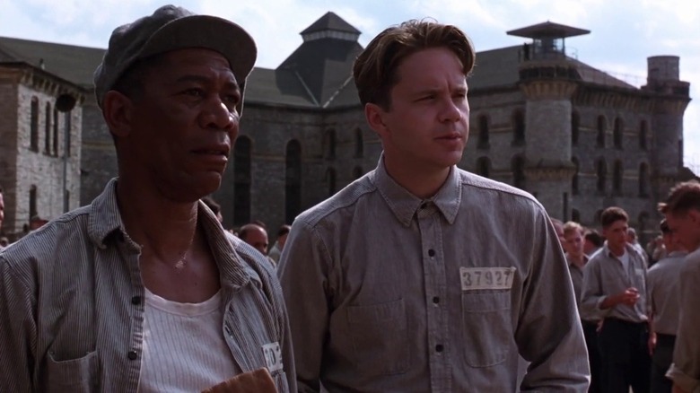 Tim Robbins as Andy Dufresne and Morgan Freeman as Red in The Shawshank Redemption, standing next to each other against the backdrop of the prison, looking on at something out of frame.