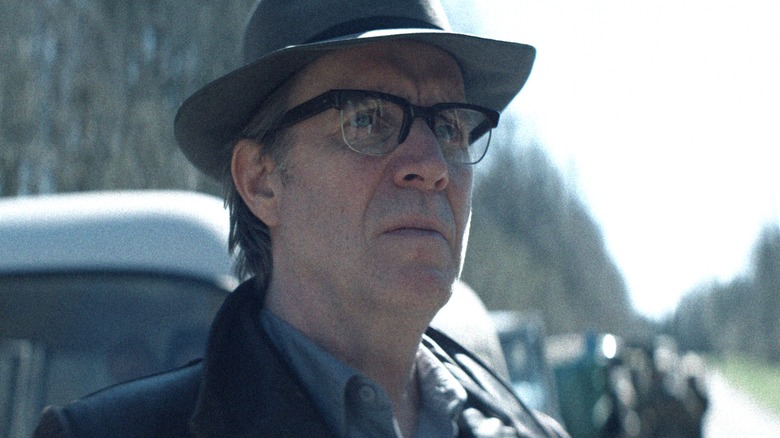 Rhys Ifans as the Chief Designer wearing a top hat and standing on a road in front of several vehicles in Star City