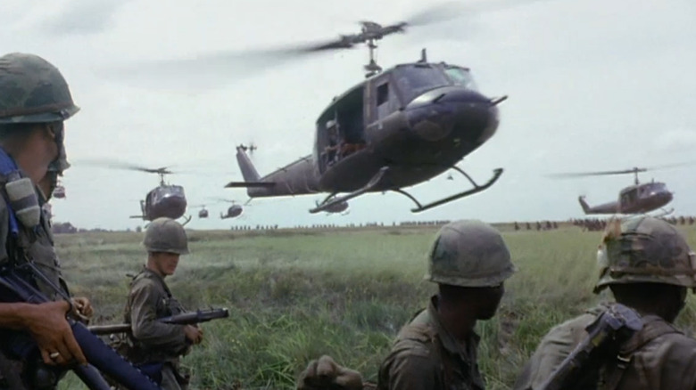 American soldiers watch helicoperters land on a grassy field in The Vietnam War