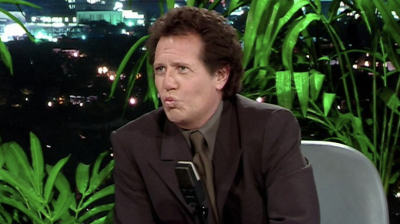 Garry Shandling as Larry Sanders sitting at his show desk in front of plants and puckering his lips in The Larry Sanders Show