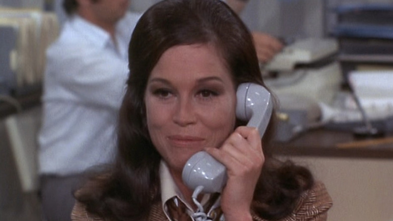 Mary Tyler Moore as Mary Richards on the phone and smiling at her desk while sitting down in The Mary Tyler Moore Show