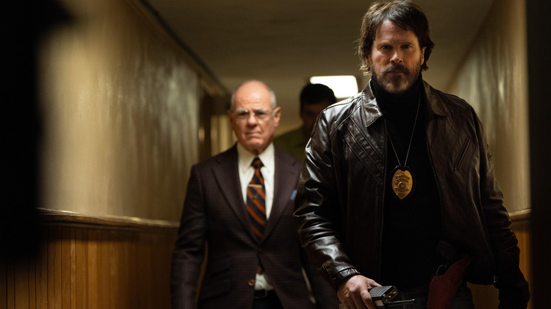 Cary Elwes as Detective Michael Grable wearing a leather jacket and walking down a hallway holding a radio followed by two others in Dead Man's Wire