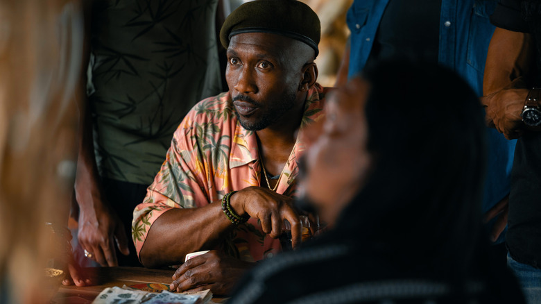 Mahershala Ali as Duncan Kincaid wearing a tropical shirt and playing a game of cards at a table in Jurassic World Rebirth