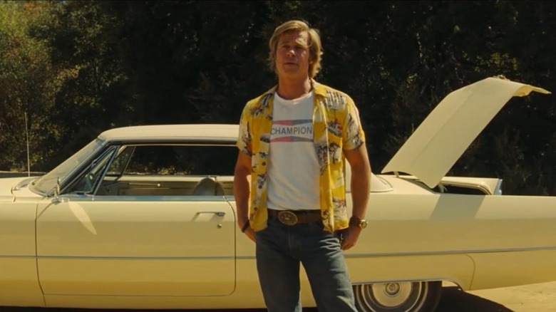 Brad Pitt as Cliff Booth, wearing his iconic Champion tee and Hawaiian shirt outfit in front of his car with the trunk popped open.